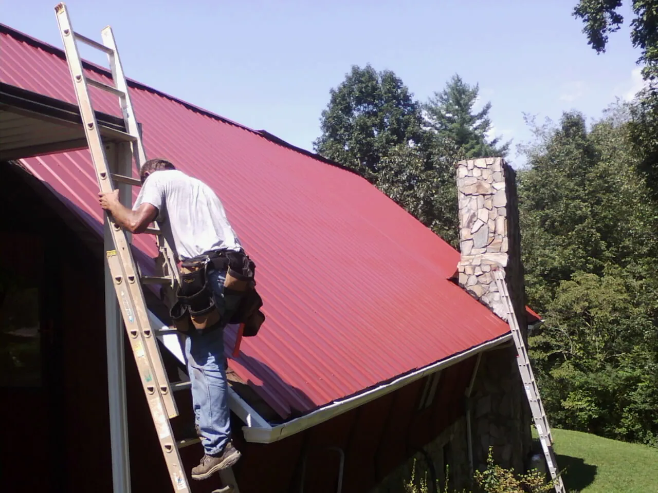 Roofer on a ladder beside a red metal roof and stone chimney — Soffit & Fascia Repair by Roofing Brothers in Hollsopple